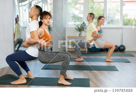 Lovers of active lifestyle perform pair yoga back to back chair pose in gym Lovers of active lifestyle perform pair yoga back to back chair pose in gym 139785888