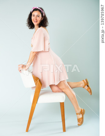 Woman in pink dress posing on chair against background of wall Woman in pink dress posing on chair against background of wall 139785967