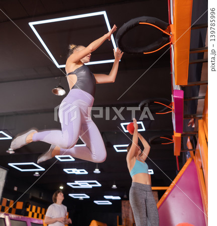 Energized woman enjoying amusement activities while jumping high on trampoline and hitting basketball hoop with ball in game club Energized woman enjoying amusement activities while jumping high on trampoline and hitting basketball hoop with ball in game club 139785996