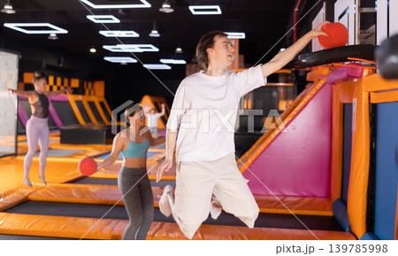 Energized young man enjoying amusement activities while jumping high on trampoline and hitting basketball hoop with ball in game club Energized young man enjoying amusement activities while jumping high on trampoline and hitting basketball hoop with ball in game club 139785998