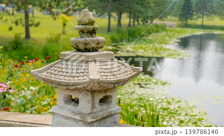 Stone lantern with intricate design beside a tranquil pond surrounded by lush greenery and blooming flowers in a serene garden setting 139786140
