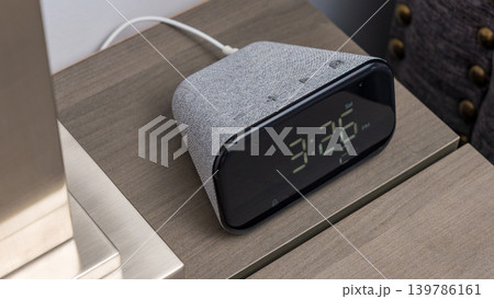 Smart home digital alarm clock with fabric finish displaying time on a bedside table next to a lamp in a modern bedroom setting 139786161