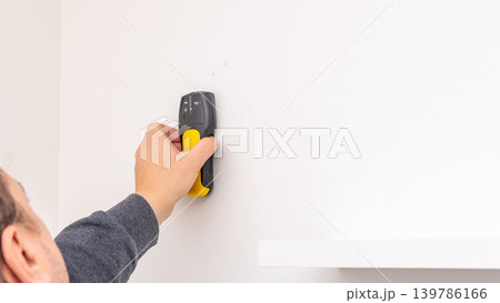 Mans hand pressing yellow stud finder on white wall above installed black shelf bracket. Home improvement, verifying wall support, DIY post-installation check, tool usage. Mans hand pressing yellow stud finder on white wall above installed black shelf bracket. Home improvement, verifying wall support, DIY post-installation check, tool usage. 139786166