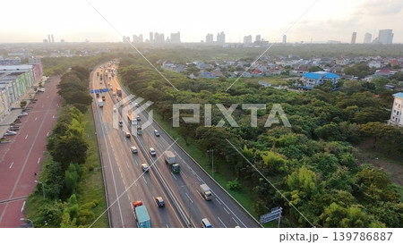 Elevated aerial view of traffic on a multi-lane highway surrounded by lush green trees, leading towards a distant city skyline at sunset. Elevated aerial view of traffic on a multi-lane highway surrounded by lush green trees, leading towards a distant city skyline at sunset. 139786887