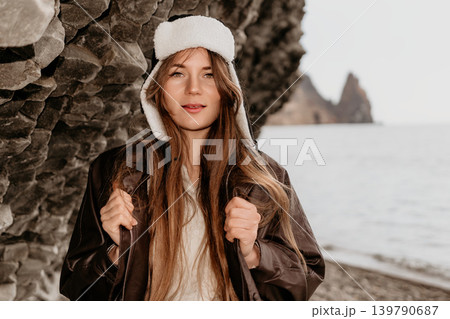 Woman ushanka coastline posing against a textured rock wall by the sea wearing a leather jacket and winter hat Woman ushanka coastline posing against a textured rock wall by the sea wearing a leather jacket and winter hat 139790687