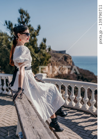 Woman cliff ocean elegant lady in white dress sitting on wooden bench overlooking a scenic coast during a sunny vacation 139790711