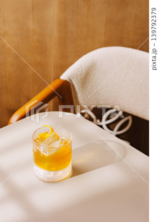 Classic cocktail with orange zest and ice cube on a table near a chair in a cozy indoor setting 139792379