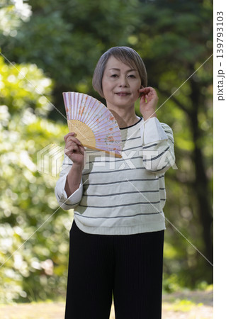新緑の公園で扇子で扇いで涼むシニア女性 新緑の公園で扇子で扇いで涼むシニア女性 139793103
