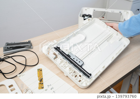 Hands working on a disassembled flatbed scanner laid out on a light wooden desk, exposing internal parts, ribbon cable and circuit board during maintenance or DIY electronics repair in a home office. 139793760