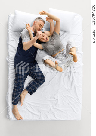 Laughing couple moving on white bed from overhead view while woman holds tablet with feet. Laughing couple moving on white bed from overhead view while woman holds tablet with feet. 139794820