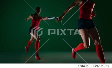 Two female runners passing relay baton in dramatic green studio scene. 139795025