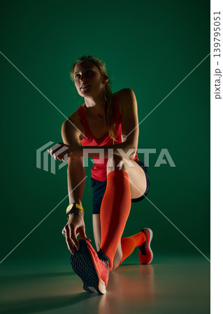 Female runner kneeling in deep stretch before training on green studio background. 139795051