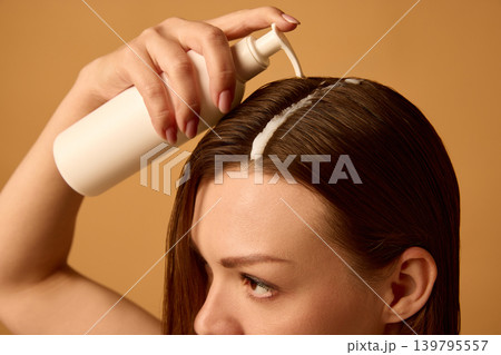 Woman applying hair serum from pump bottle to wet brunette hair on warm studio background. 139795557