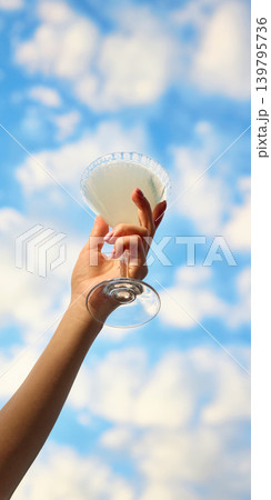 Elegant white cocktail in coupe glass raised against blue sky. Elegant white cocktail in coupe glass raised against blue sky. 139795736