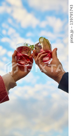 Two red cocktails with cucumber garnish raised toward blue sky. Two red cocktails with cucumber garnish raised toward blue sky. 139795743