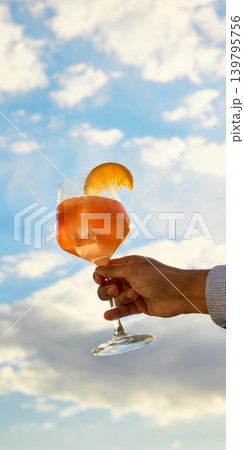 Orange cocktail glass glowing against blue sky and clouds. Orange cocktail glass glowing against blue sky and clouds. 139795756