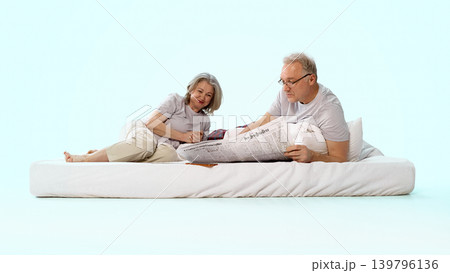 Older couple relaxing on bed while man reads newspaper in pastel interior. Older couple relaxing on bed while man reads newspaper in pastel interior. 139796136