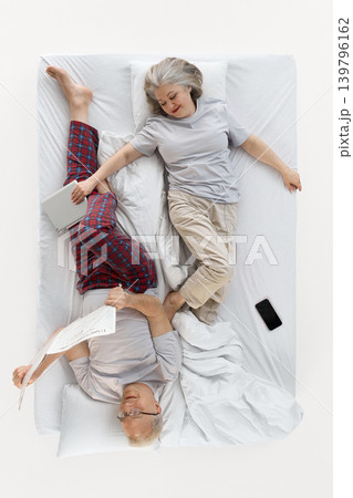 Older woman resting beside man reading newspaper in white bed top view. Older woman resting beside man reading newspaper in white bed top view. 139796162
