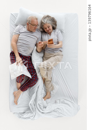 Senior woman showing smartphone to man while resting together in white bed. Senior woman showing smartphone to man while resting together in white bed. 139796164