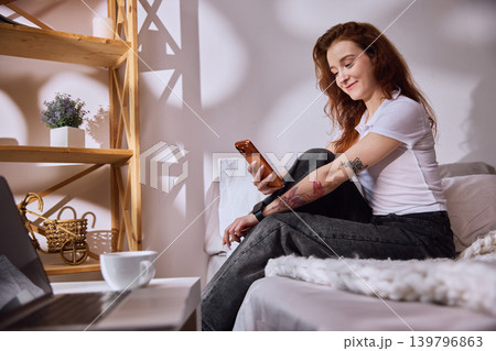 Red haired woman checking smartphone on bed near laptop. Red haired woman checking smartphone on bed near laptop. 139796863