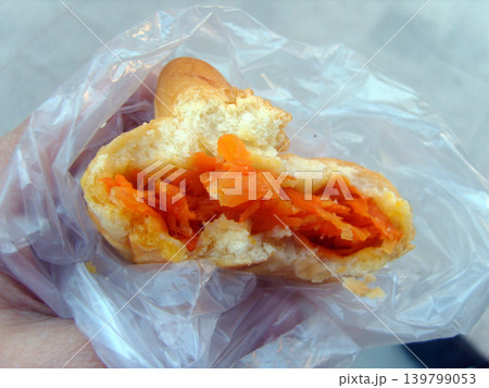Broken baked bun filled with grated orange carrots, held in a hand inside a clear plastic bag. The soft pastry showcases a moist, vibrant filling, typical of traditional street food snacks Broken baked bun filled with grated orange carrots, held in a hand inside a clear plastic bag. The soft pastry showcases a moist, vibrant filling, typical of traditional street food snacks 139799053