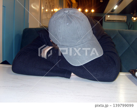 Tired person in a grey baseball cap and black jacket sleeps with their head resting on folded arms on a white table inside a cafe. Tired person in a grey baseball cap and black jacket sleeps with their head resting on folded arms on a white table inside a cafe. 139799099
