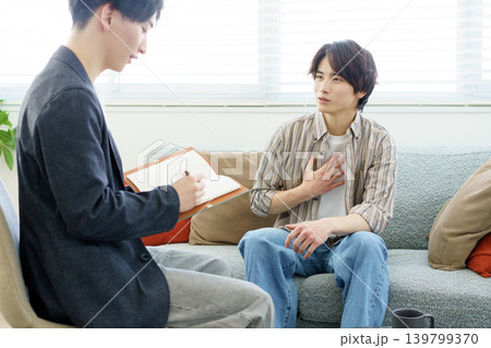カウンセリング ソファに座って話をする若い日本人男性と話を聞きながらメモをする若い日本人医師 カウンセリング ソファに座って話をする若い日本人男性と話を聞きながらメモをする若い日本人医師 139799370