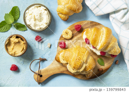 French pastry, freshly baked croissants with peanut butter, raspberries and banana on a blue stone background. Healthy food dessert concept. View from above. French pastry, freshly baked croissants with peanut butter, raspberries and banana on a blue stone background. Healthy food dessert concept. View from above. 139799843