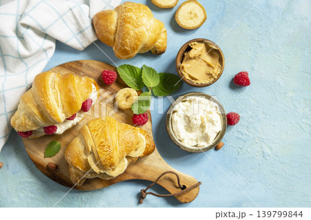 French pastry, freshly baked croissants with peanut butter, raspberries and banana on a blue stone background. Healthy food dessert concept. View from above. Copy space. French pastry, freshly baked croissants with peanut butter, raspberries and banana on a blue stone background. Healthy food dessert concept. View from above. Copy space. 139799844