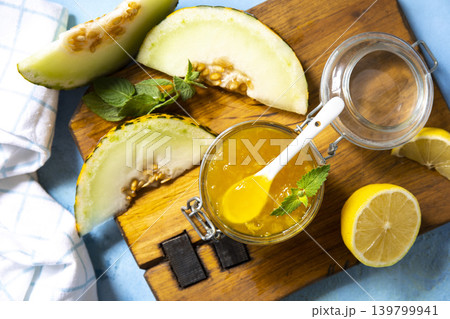 Sweet melon and citrus jam, homemade jelly preserves in small glass jar with fresh melon slices on a blue stone tabletop. View from above. 139799941