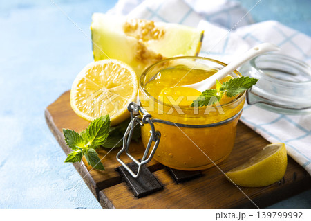 Sweet melon and citrus jam, homemade jelly preserves in small glass jar with fresh melon slices on a blue stone tabletop. Homemade preserve. Copy space. 139799992