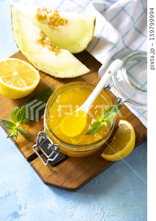 Sweet melon and citrus jam, homemade jelly preserves in small glass jar with fresh melon slices on a blue stone tabletop. Homemade preserve. Copy space. 139799994