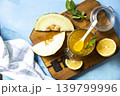 Sweet melon and citrus jam, homemade jelly preserves in small glass jar with fresh melon slices on a blue stone tabletop. Homemade preserve. View from above. 139799996