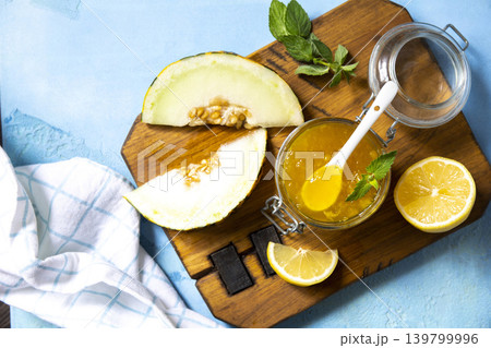 Sweet melon and citrus jam, homemade jelly preserves in small glass jar with fresh melon slices on a blue stone tabletop. Homemade preserve. View from above. Sweet melon and citrus jam, homemade jelly preserves in small glass jar with fresh melon slices on a blue stone tabletop. Homemade preserve. View from above. 139799996