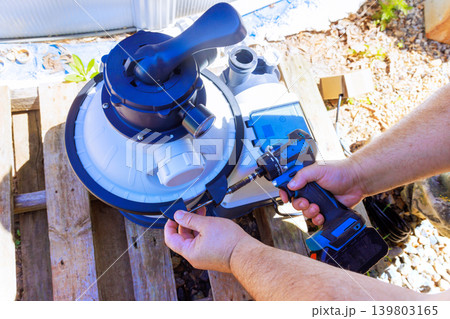 Worker uses drill to fix pool filter system while standing in backyard during day. Worker uses drill to fix pool filter system while standing in backyard during day. 139803165