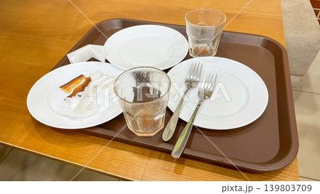 A brown plastic tray with dirty empty dishes, used glasses, forks, and food leftovers on a wooden table after a meal 139803709