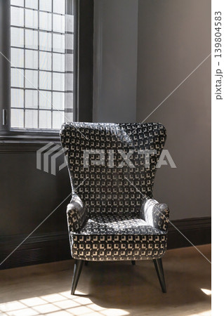 A stately wingback armchair rests gracefully in a sunlit gallery alcove. Its bold upholstery features a precise, geometric tapestry of repeating monochrome squares. Streams of soft daylight pour A stately wingback armchair rests gracefully in a sunlit gallery alcove. Its bold upholstery features a precise, geometric tapestry of repeating monochrome squares. Streams of soft daylight pour 139804583