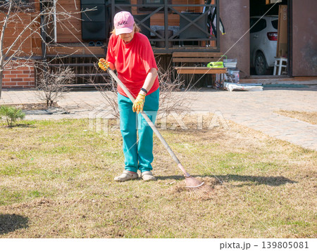 Senior woman raking dry grass in a home lawn. Gardener working in the backyard during spring season. Seasonal yard maintenance, cleaning garden soil, and landscape care activity concept. 139805081