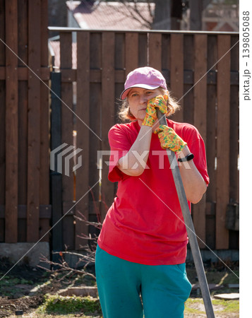 Mature woman gardener in red t-shirt leaning on garden tool in backyard. Caucasian lady in baseball cap working outdoors. Agriculture, vegetable garden maintenance and hobby lifestyle concept. Mature woman gardener in red t-shirt leaning on garden tool in backyard. Caucasian lady in baseball cap working outdoors. Agriculture, vegetable garden maintenance and hobby lifestyle concept. 139805088