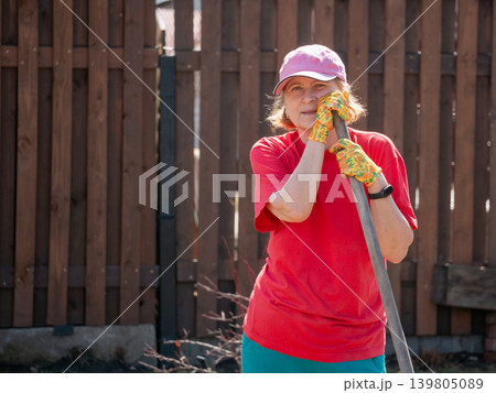 Senior woman gardener leaning on spade in backyard. Mature lady wearing cap and work glove doing yard work. Hobby of horticulture and vegetable growing in spring season near house fence. Senior woman gardener leaning on spade in backyard. Mature lady wearing cap and work glove doing yard work. Hobby of horticulture and vegetable growing in spring season near house fence. 139805089