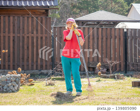 Woman gardener resting with rake in backyard. Mature lady cleaning lawn in spring season. Housework and yard maintenance. Gardening concept, lifestyle activity at home property. Woman gardener resting with rake in backyard. Mature lady cleaning lawn in spring season. Housework and yard maintenance. Gardening concept, lifestyle activity at home property. 139805090