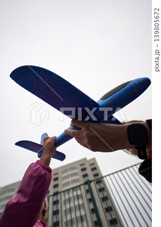 Young child lifts foam glider toward sky. Child with family in city outdoors scene playing involving foam glider Young child lifts foam glider toward sky. Child with family in city outdoors scene playing involving foam glider 139805672