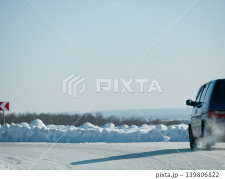 Blue car driving on a white snowy road with a white snow bank. Winter travel and cold weather condition. Season background. Blue car driving on a white snowy road with a white snow bank. Winter travel and cold weather condition. Season background. 139806822