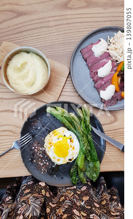 Woman eating delicious meal with roasted beef, fried egg, lettuce and mashed potato at restaurant. Gourmet dinner set served on plate. Top view of healthy food culture and dining. 139807055