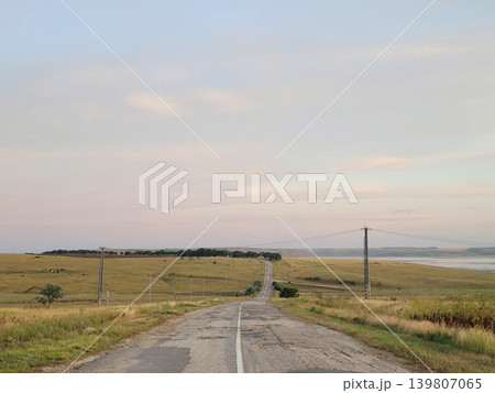 Empty asphalt road stretching through grassy field toward horizon during sunset. Rural country landscape with power line and lake in distance. Travel, journey and nature exploration concept. Empty asphalt road stretching through grassy field toward horizon during sunset. Rural country landscape with power line and lake in distance. Travel, journey and nature exploration concept. 139807065