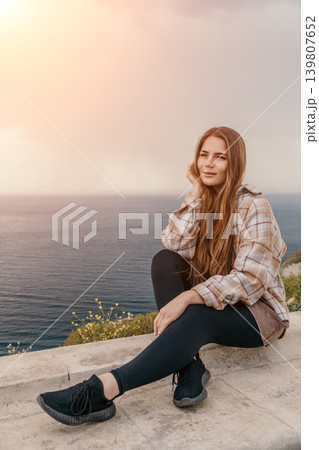 Woman sea sunset sitting on stone ledge overlooking Mediterranean coastal view with copy space and beautiful sky Woman sea sunset sitting on stone ledge overlooking Mediterranean coastal view with copy space and beautiful sky 139807652