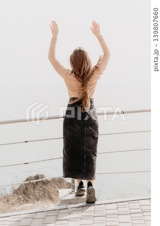 Woman viewpoint freedom. Young lady standing at a coastal balcony with her hands raised towards the bright sky. Woman viewpoint freedom. Young lady standing at a coastal balcony with her hands raised towards the bright sky. 139807660