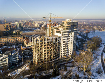Apartment or office tall building under construction, aerial view. Tower crane silhouette Snowy field and distant city on bright blue sky copy space background. Drone photography. Apartment or office tall building under construction, aerial view. Tower crane silhouette Snowy field and distant city on bright blue sky copy space background. Drone photography. 139807894