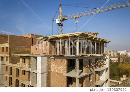 Apartment or office tall building under construction, top view. Brick walls, scaffolding and concrete support pillars. Tower crane on bright blue sky copy space background. Drone aerial photography. Apartment or office tall building under construction, top view. Brick walls, scaffolding and concrete support pillars. Tower crane on bright blue sky copy space background. Drone aerial photography. 139807922
