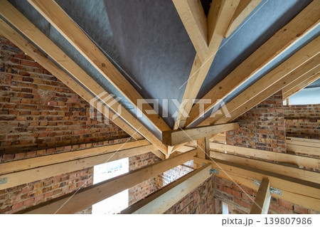 Attic space of a building under construction with wooden beams of a roof structure and brick walls. Real estate development concept. Attic space of a building under construction with wooden beams of a roof structure and brick walls. Real estate development concept. 139807986